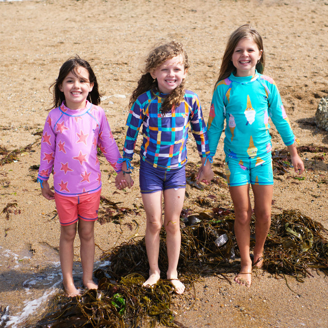 Three girls in UV Protective Surf Sets Multi Check, starfish and ice cream designs stand on a beach holding hands, smiling at the camera.